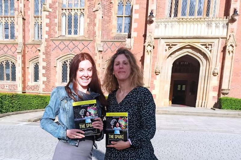 two smiling women holding a copy each of a dark-cover book with an old red brick building and doorway behind